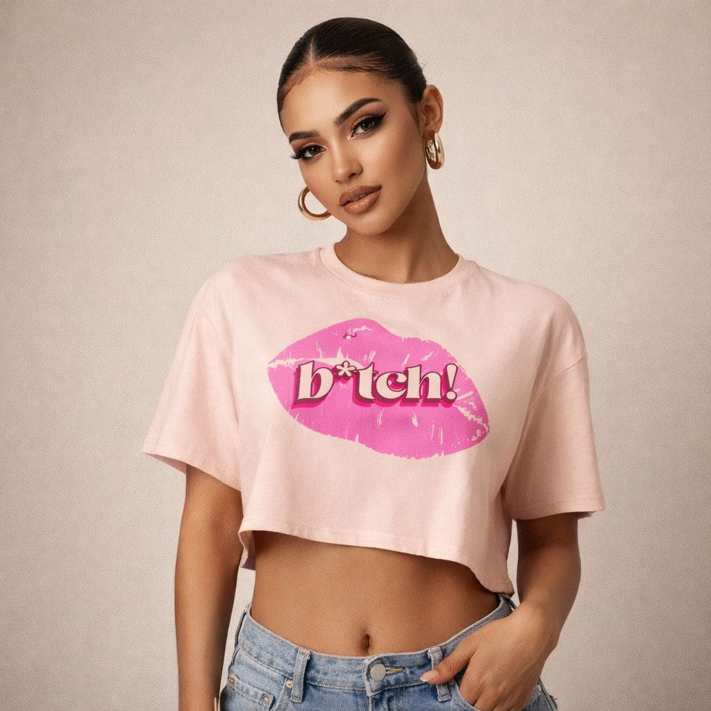A woman wearing hoop earrings and a light pink crop top with a large pink lip print and the word "b*tch!" printed on it, paired with blue jeans, standing against a neutral background.