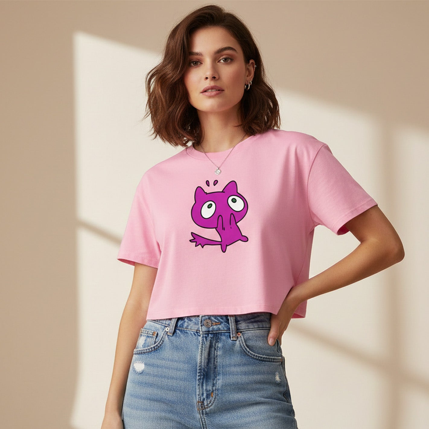 A woman wearing a pink cropped t-shirt with a cartoonish purple cat graphic and high-waisted blue jeans stands in a softly lit room with a beige background.