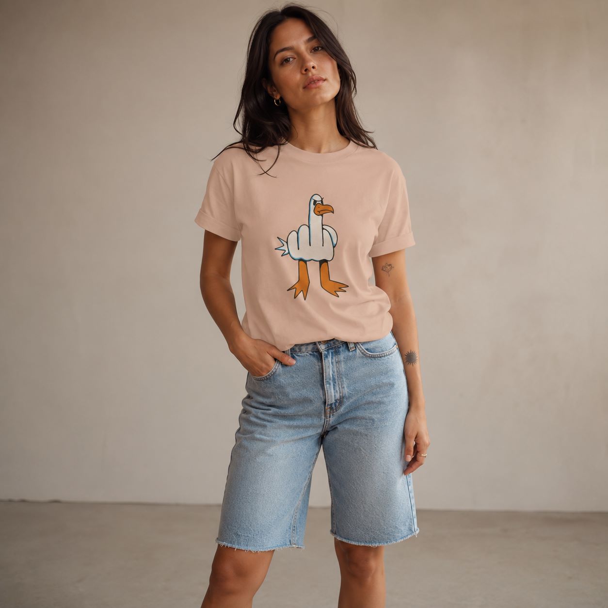 A woman modeling a light peach-pink graphic t-shirt with a cartoon duck design, paired with light-wash denim shorts, standing against a neutral beige wall.