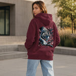 A woman wearing a burgundy hoodie with a large graphic of a tiger and cherry blossoms on the back, paired with light blue jeans, standing outdoors in front of a modern building.