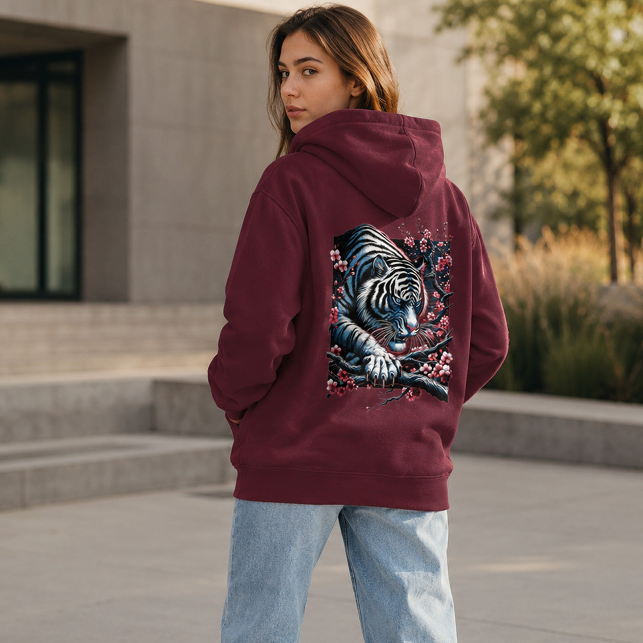 A woman wearing a burgundy hoodie with a large graphic of a tiger and cherry blossoms on the back, paired with light blue jeans, standing outdoors in front of a modern building.
