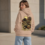 A woman wearing a beige hoodie with a large, detailed multicolor graphic print on the back, shown from behind in an outdoor setting.