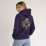 A woman wearing a purple hoodie with a large graphic print on the back, featuring a woman with long hair, crows, and wolves. She is wearing light blue jeans and posing against a plain light gray background.