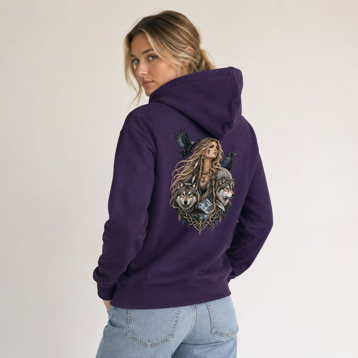 A woman wearing a purple hoodie with a large graphic print on the back, featuring a woman with long hair, crows, and wolves. She is wearing light blue jeans and posing against a plain light gray background.
