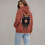 A woman wearing a terracotta-colored hoodie with a large graphic of a tiger and a woman's face on the back, paired with light blue jeans, photographed against a neutral gray background.