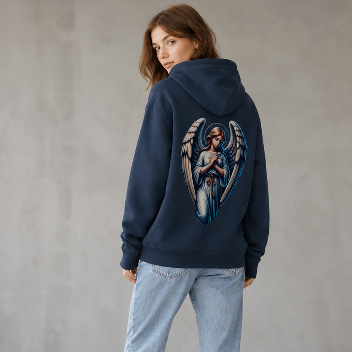 A woman wearing a navy blue hoodie with a large graphic of an angel on the back, paired with light blue jeans, standing against a textured gray wall.