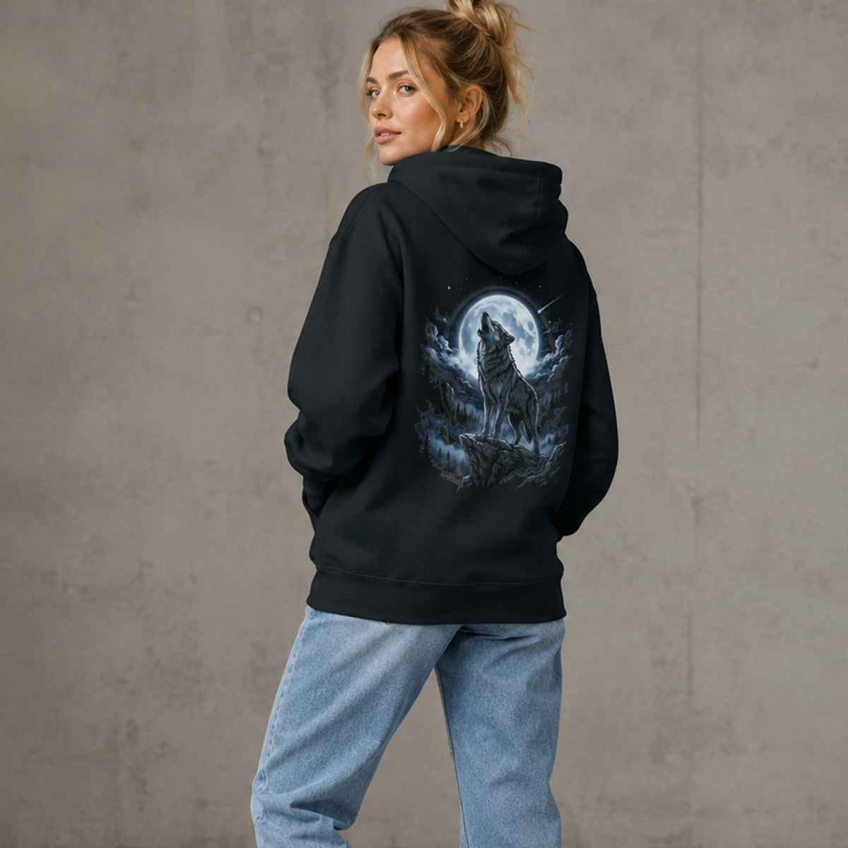 A woman wearing a black hoodie with a large wolf howling at a full moon graphic on the back, paired with light blue jeans, standing against a textured gray concrete wall.