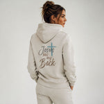 A woman shown from the back wearing a light beige hoodie with the text 'Jesus has my Back' and a blue cross graphic printed on the back, paired with matching beige pants against a plain light gray studio background.