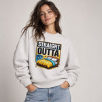 A woman wearing a light gray crewneck sweatshirt with a colorful graphic print that reads 'STRAIGHT OUTTA' above an illustration of a bed with yellow and blue bedding. She is paired with light wash blue jeans against a neutral light gray studio background.