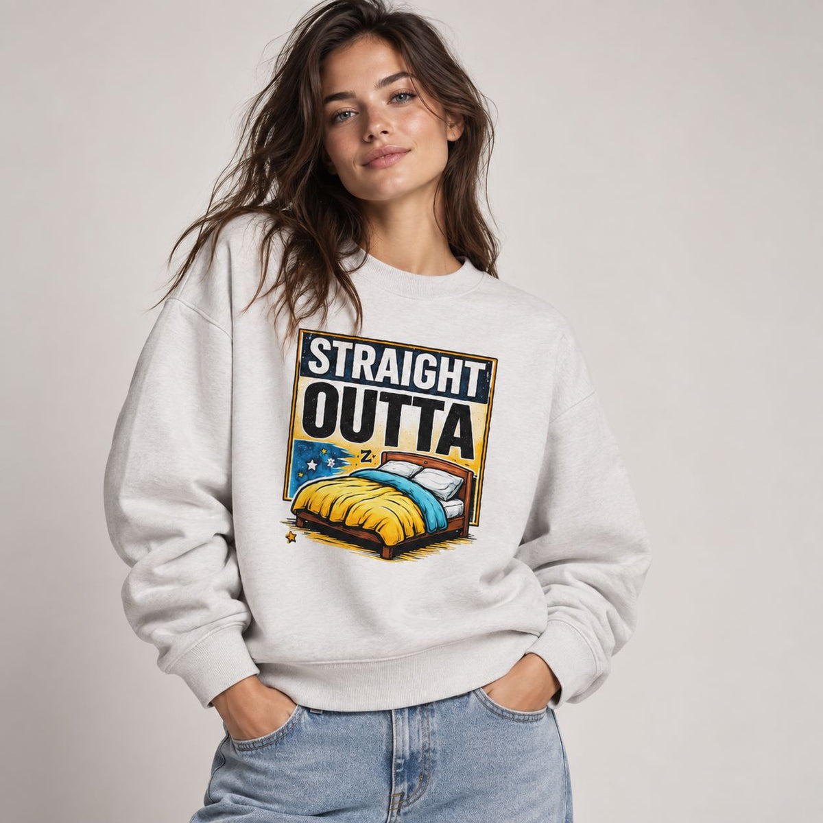 A woman wearing a light gray crewneck sweatshirt with a colorful graphic print that reads 'STRAIGHT OUTTA' above an illustration of a bed with yellow and blue bedding. She is paired with light wash blue jeans against a neutral light gray studio background.