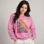 A woman wearing a pink crewneck sweatshirt with a colorful retro-style text graphic that reads 'I AM NOT PERFECT BUT I AM LIMITED EDITION'. She is posing with her hands in her white pants against a solid light gray background.