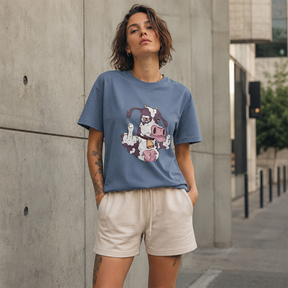 A woman wearing a dusty blue graphic t-shirt with a cow illustration and cream-colored shorts, standing outdoors against a concrete wall.
