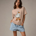 A woman modeling a beige graphic t-shirt with a cartoon goose giving a middle finger, paired with light blue denim shorts, against a solid neutral studio background.