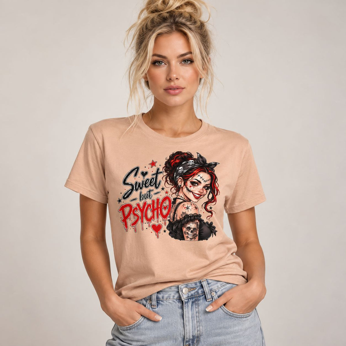 A woman wearing a light peach graphic t-shirt with a gothic 'Sweet but Psycho' design, paired with light-wash blue jeans, set against a plain light gray studio background.