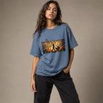 A woman modeling an oversized blue graphic t-shirt featuring 'THE WALKING DEAD' and 'MIA' in a fiery orange and black print, paired with dark gray pants, against a solid beige studio background.