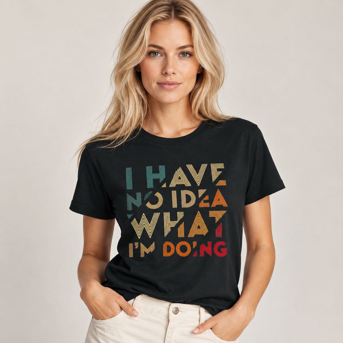 A woman wearing a black graphic t-shirt with the text 'I HAVE NO IDEA WHAT I'M DOING' in a retro, multi-colored font, paired with off-white pants, set against a plain light gray background.