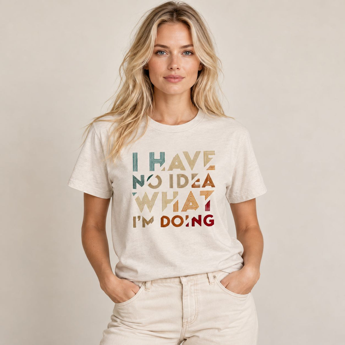 A woman wearing a light beige graphic t-shirt with the text 'I HAVE NO IDEA WHAT I'M DOING' in a retro, multi-colored block pattern, paired with light beige jeans, against a plain beige background.