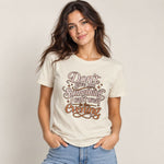 A woman wearing a cream-colored graphic t-shirt with brown and gold text that reads 'Don't give me Something I can't wear in the Evening', paired with light blue jeans, against a plain beige background.