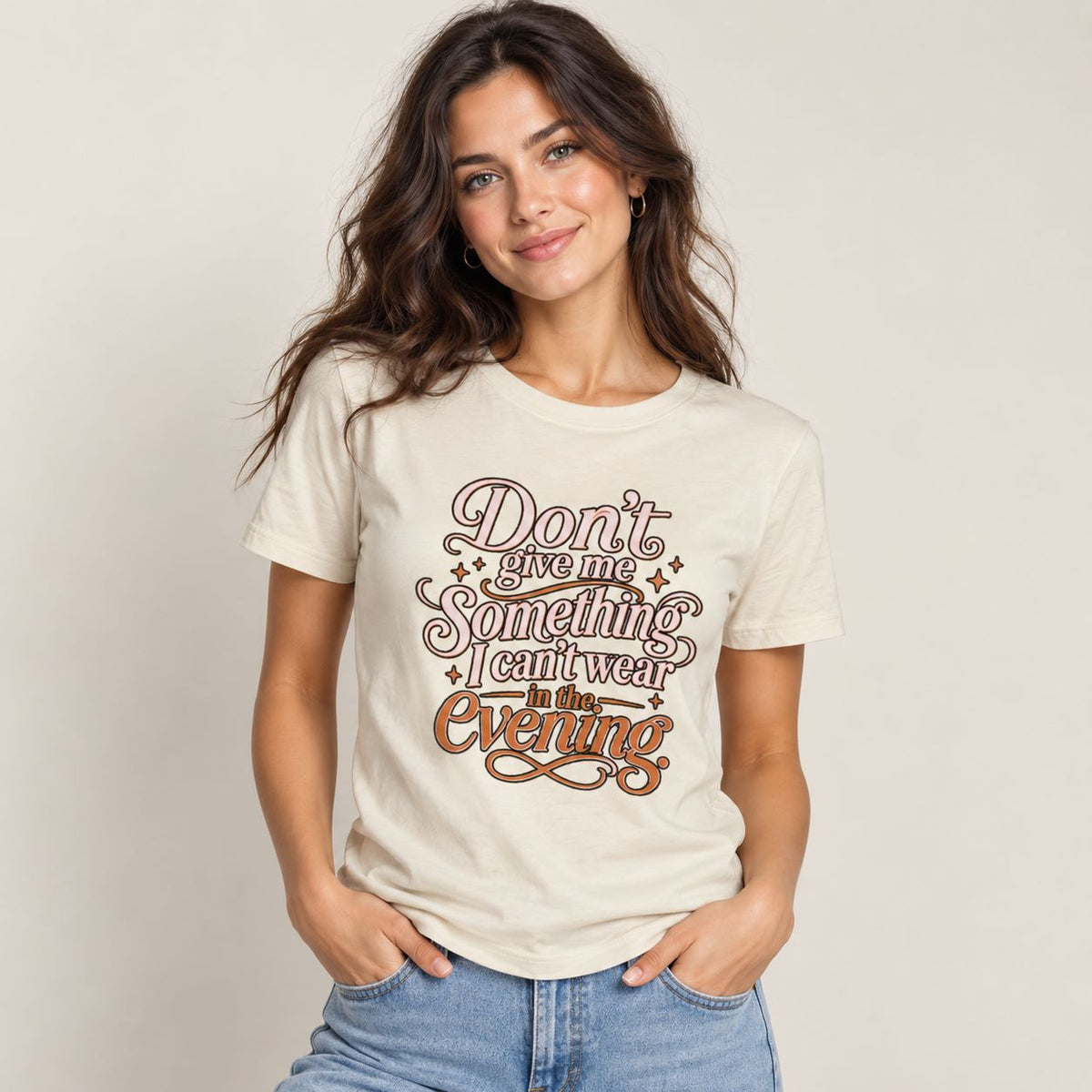 A woman wearing a cream-colored graphic t-shirt with brown and gold text that reads 'Don't give me Something I can't wear in the Evening', paired with light blue jeans, against a plain beige background.