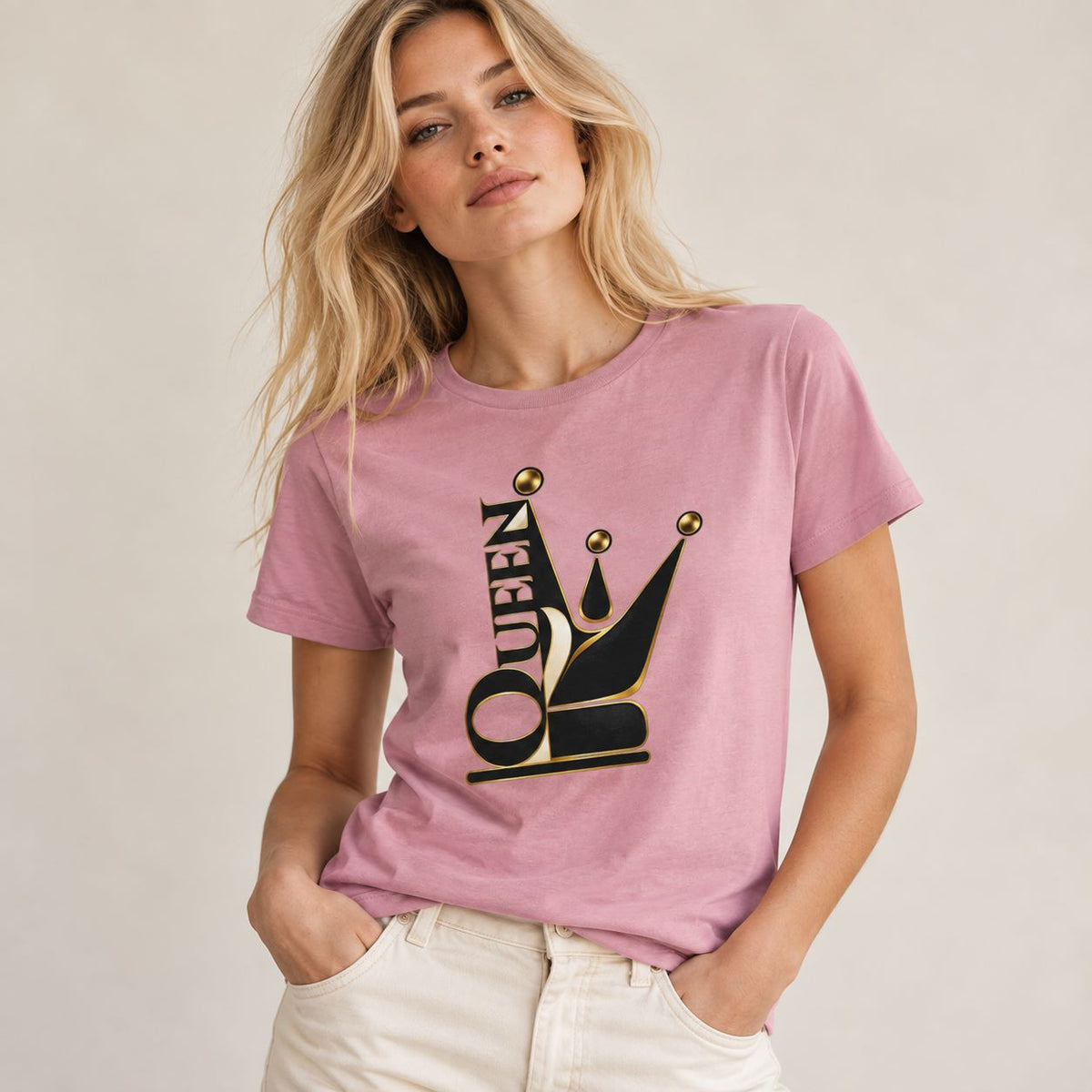 A woman wearing a dusty pink short-sleeve t-shirt with a black and gold crown graphic and the word 'QUEEN' integrated into it, paired with off-white pants, against a neutral beige background.