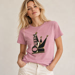 A woman wearing a dusty pink short-sleeve t-shirt with a black and gold crown graphic and the word 'QUEEN' integrated into it, paired with off-white pants, against a neutral beige background.