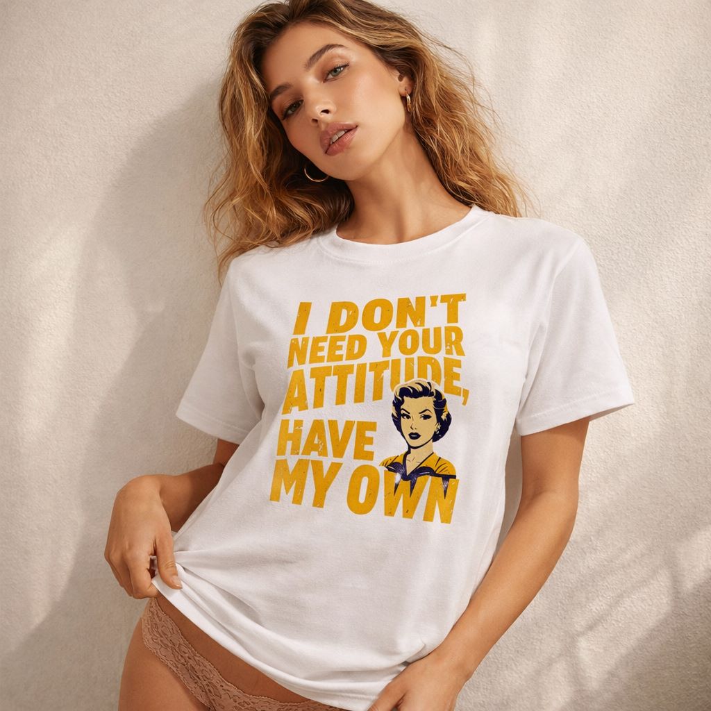 A woman with long, wavy hair wears a white T-shirt with the text "I DON'T NEED YOUR ATTITUDE, HAVE MY OWN" and an illustrated woman's face. She poses indoors, holding the bottom of the shirt and wearing lace underwear.