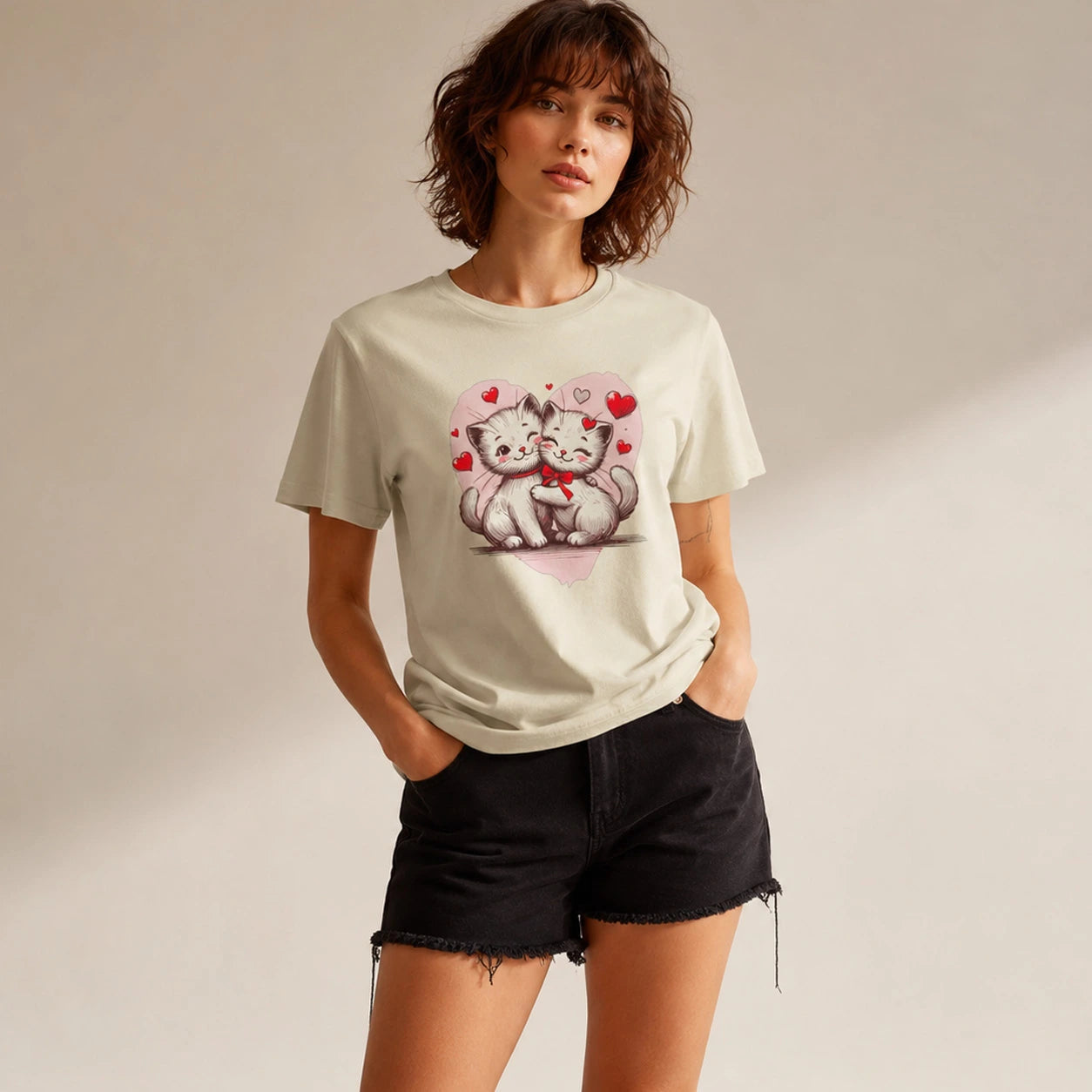 A woman modeling a light beige graphic t-shirt with a cute cartoon cat design, paired with black denim shorts, set against a warm neutral studio background.