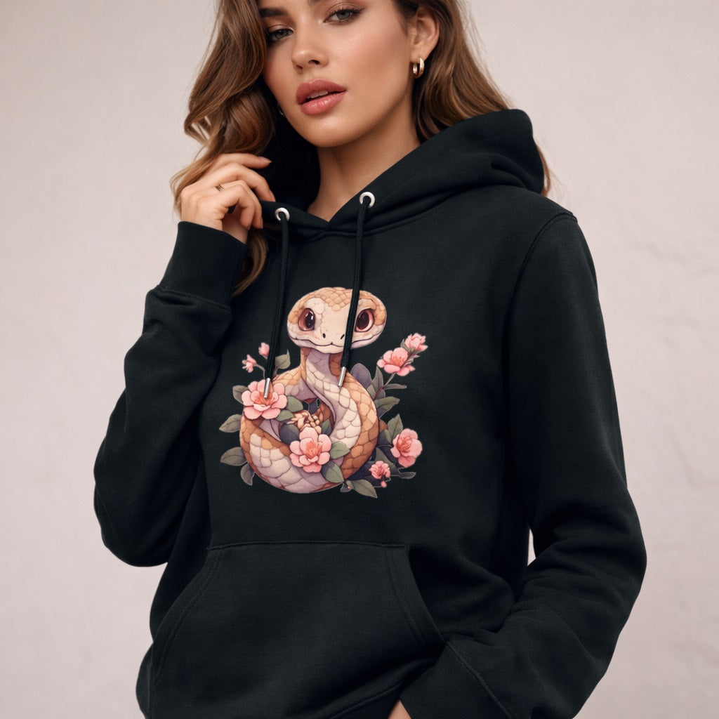 A woman wearing a black hoodie featuring a cute cartoon snake surrounded by pink flowers. She has wavy brown hair, hoop earrings, and is standing against a light background.