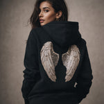 Woman wearing a black hoodie with angel wings design on the back against a neutral background