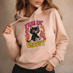 A woman wearing a light pink hoodie with a colorful graphic of a black cat wearing star-shaped glasses, surrounded by vibrant swirls, and the words “Feelin’ Groovy” printed on the front.