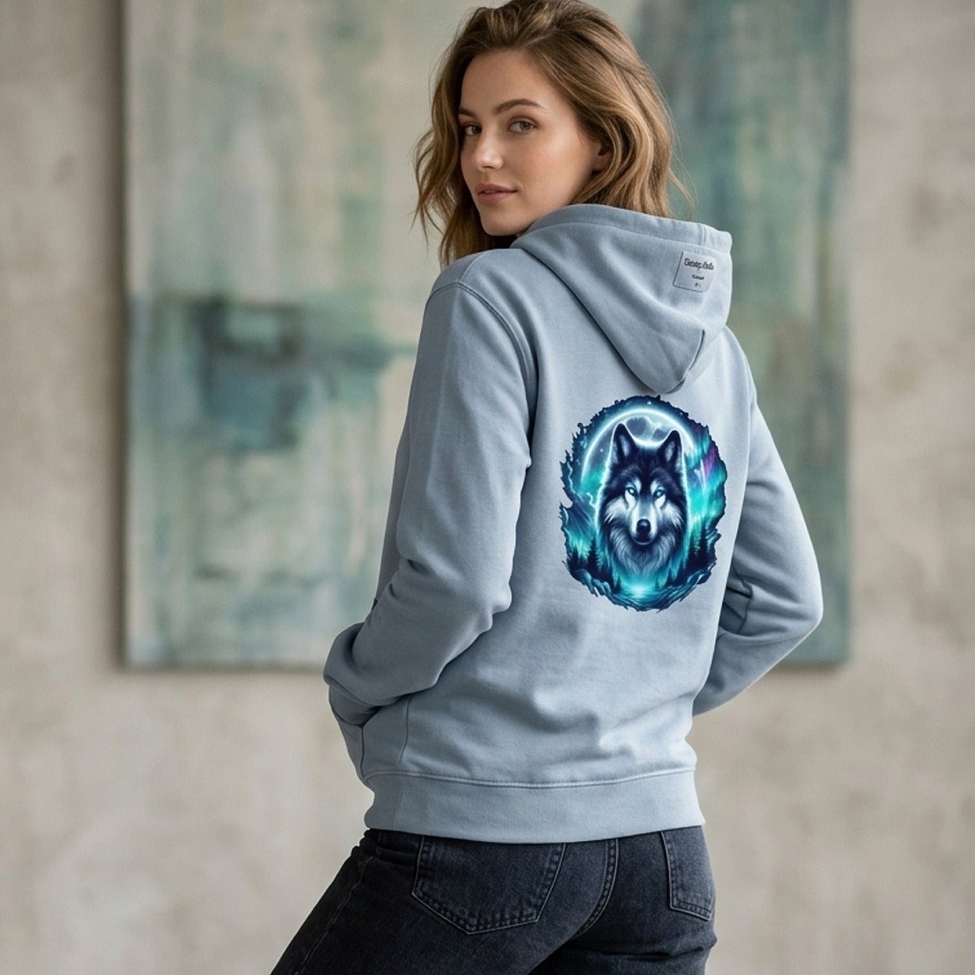 Woman wearing a light blue hoodie with a wolf design on the back, standing in front of a painting.