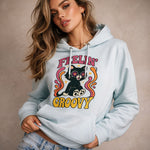 A woman wearing a light blue hoodie with a colorful graphic of a cat with starry eyes, surrounded by swirls, and the words "Feelin' Groovy." She has long wavy hair and is posing against a neutral background.
