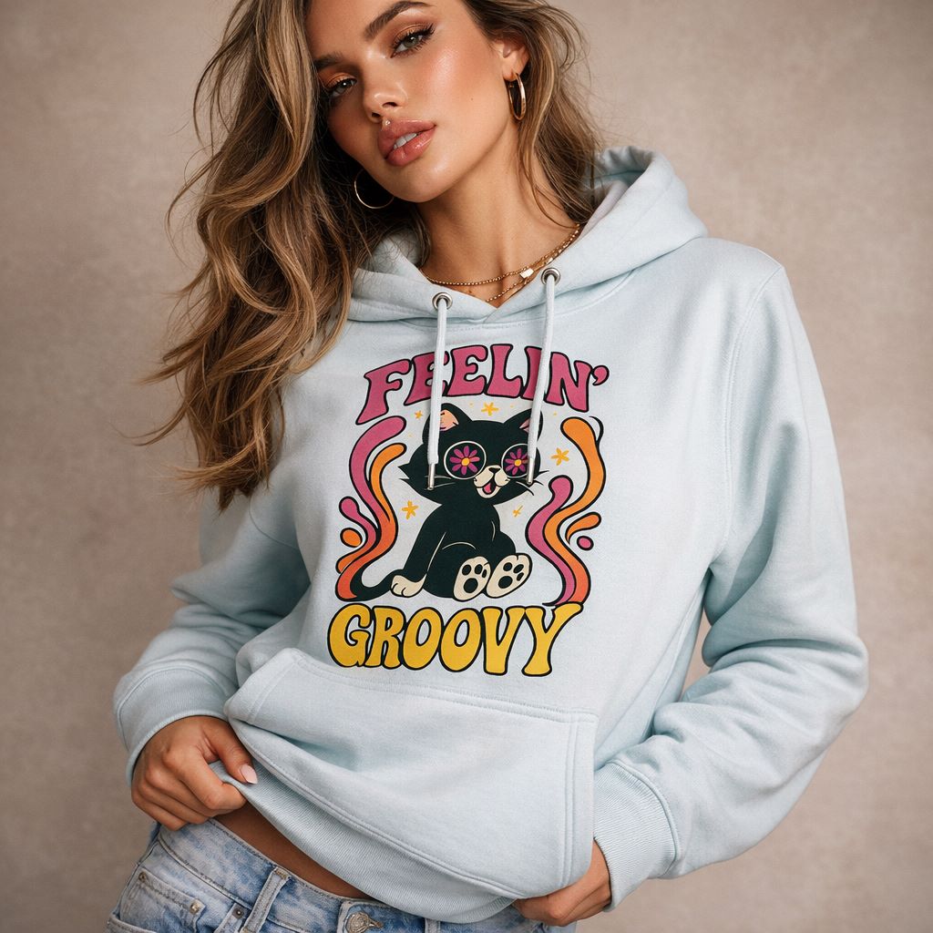 A woman wearing a light blue hoodie with a colorful graphic of a cat with starry eyes, surrounded by swirls, and the words "Feelin' Groovy." She has long wavy hair and is posing against a neutral background.