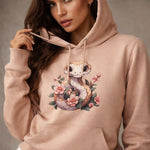 A woman wearing a light pink hoodie featuring a cute cartoon snake surrounded by pink flowers. She is holding the hood's drawstring and looking at the camera.