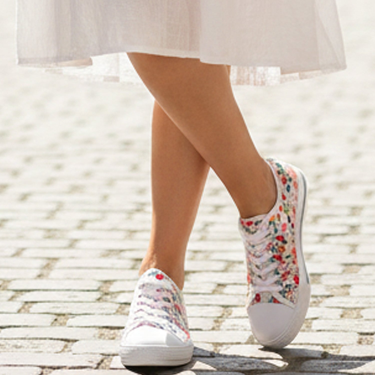White sneakers with colorful floral pattern worn by a person in a light dress on a stone pavement.