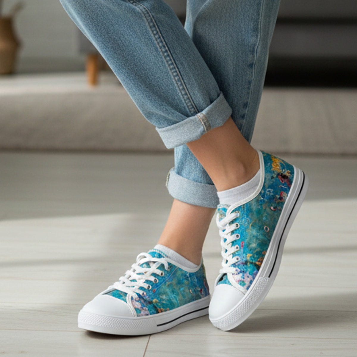 Person wearing rolled-up blue jeans and teal-blue sneakers with artistic paint splatter designs, standing on a light wooden floor in a modern, cozy room.