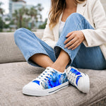 A woman in blue jeans, a white top, and a light blazer sits outdoors on a couch, crossing her legs. She is wearing white sneakers with a vibrant blue abstract design.