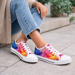 Woman wearing blue jeans and a beige sweater sits on a stone step, showing off colorful, tie-dye style sneakers with white laces and soles. Green plants are visible in the background.