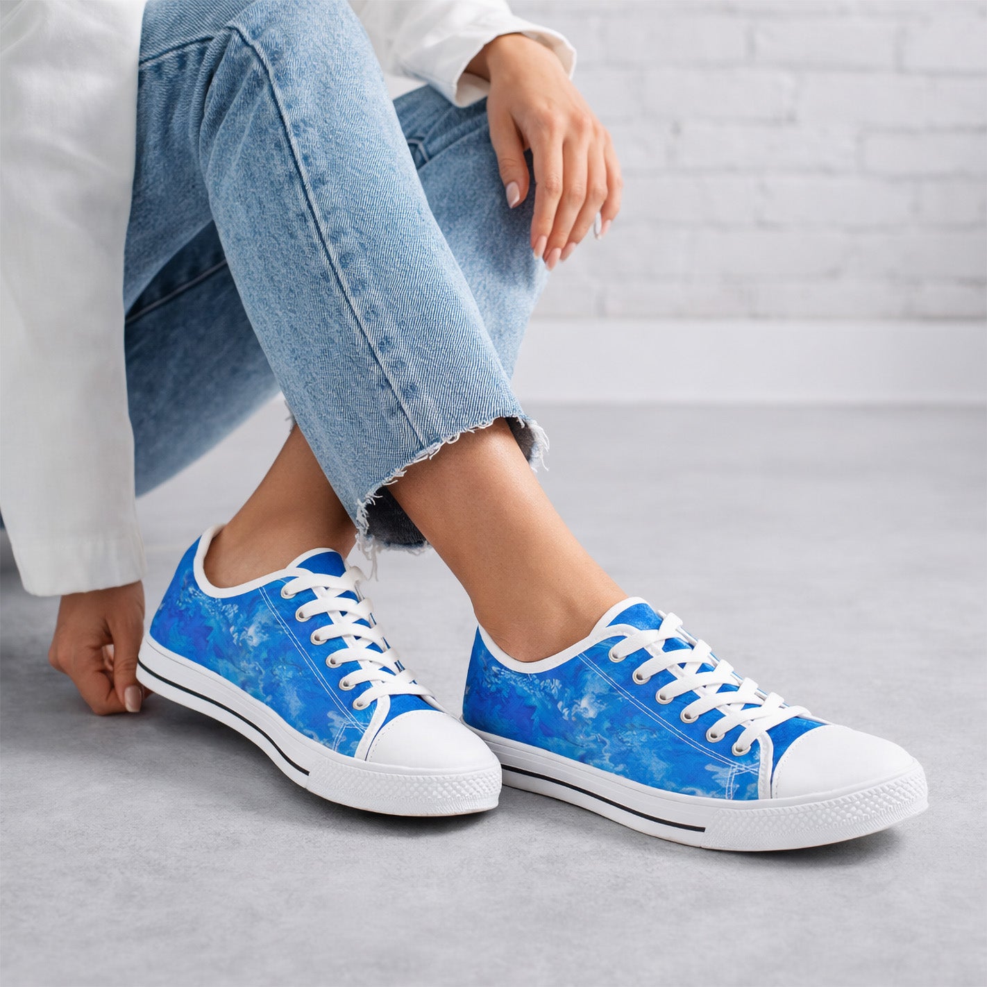 Woman wearing light blue jeans and a white shirt sits on a gray floor, sporting bright blue and white sneakers with a marbled pattern. The background features a white brick wall.