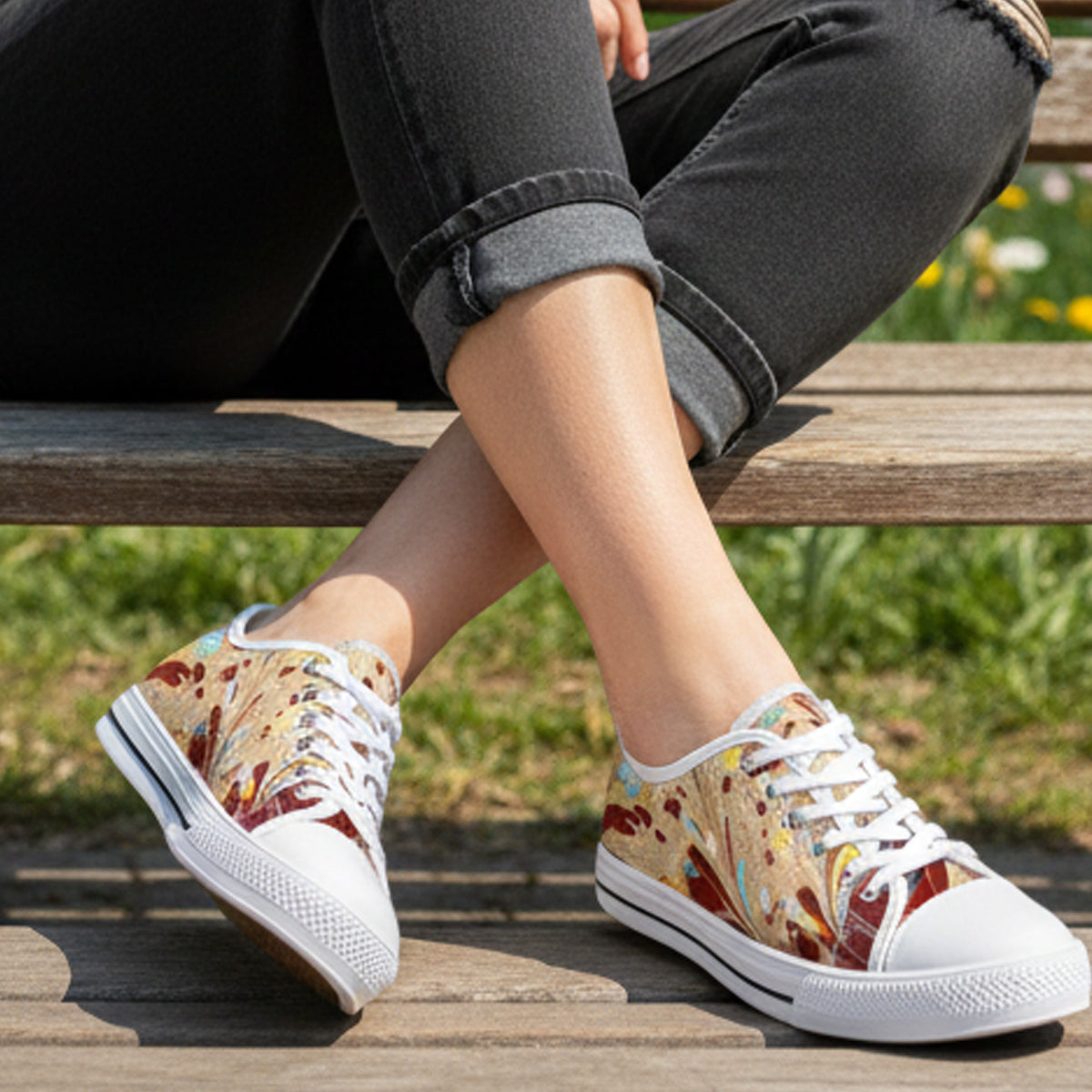 A woman wearing colorful, patterned sneakers and black rolled-up jeans sits on a wooden bench outdoors, legs crossed, with green grass and flowers in the background.