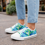 Womanwearing light blue jeans and colorful sneakers with a blue, green, and white floral pattern stands on a city sidewalk. The focus is on the shoes and lower legs.
