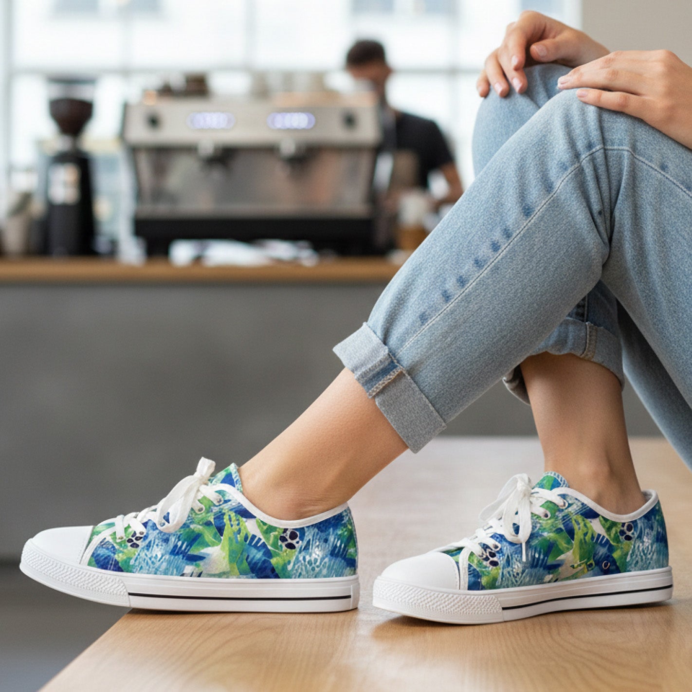 A woman wearing light blue jeans and colorful, tropical-patterned sneakers sits on a wooden counter in a café, with a coffee machine and a blurry figure in the background.