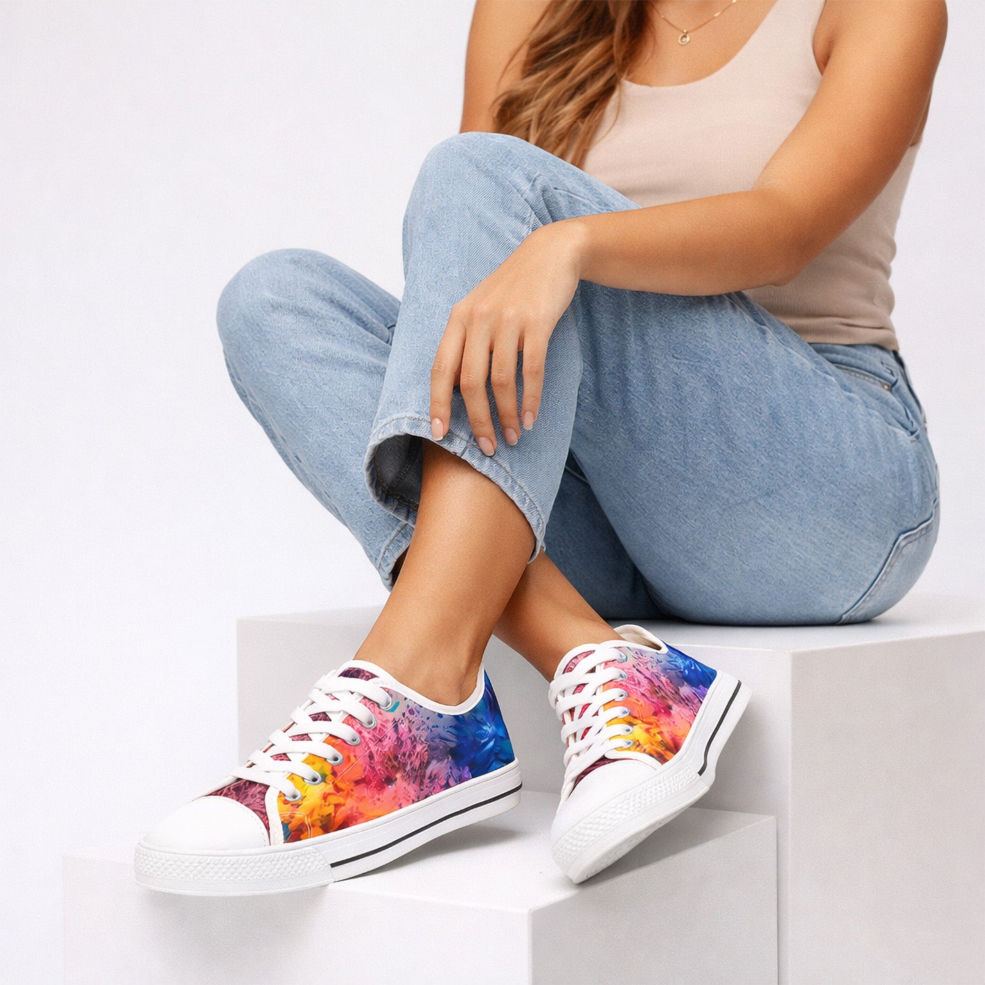 A woman wearing light blue jeans and a beige tank top sits on white steps, showing off vibrant, multicolored tie-dye sneakers with white laces and soles. Her face is not visible.