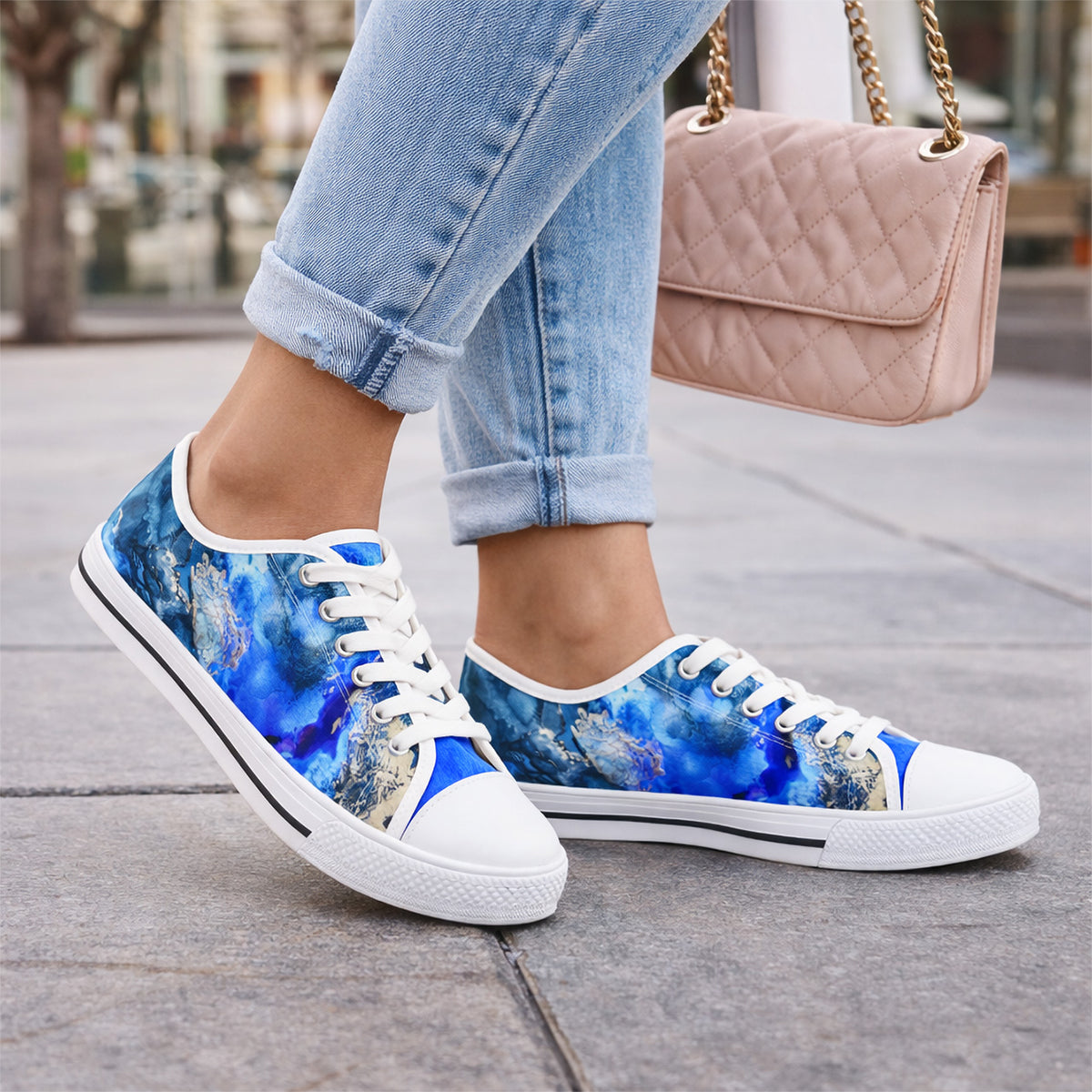 Person wearing light blue rolled-up jeans and blue cosmic-patterned sneakers, standing on a city sidewalk. They are carrying a blush pink quilted handbag with a gold chain strap.