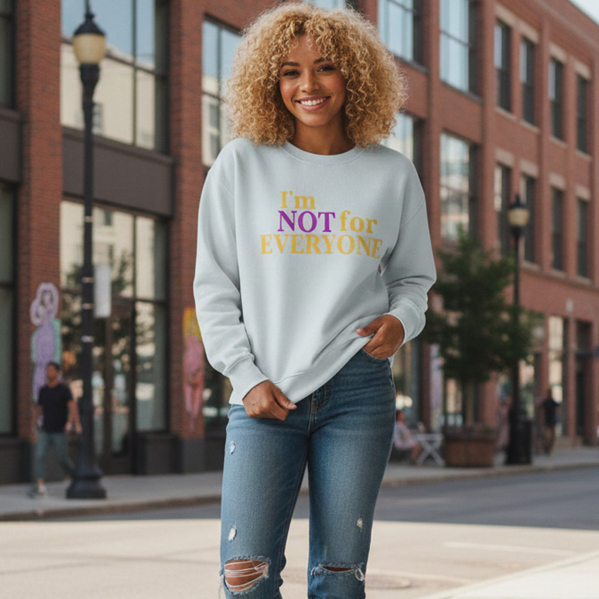 A smiling woman with curly blonde hair wears a light sweatshirt that says "I'm NOT for EVERYONE" in purple and yellow letters, standing on a city sidewalk with brick buildings and a streetlamp in the background.