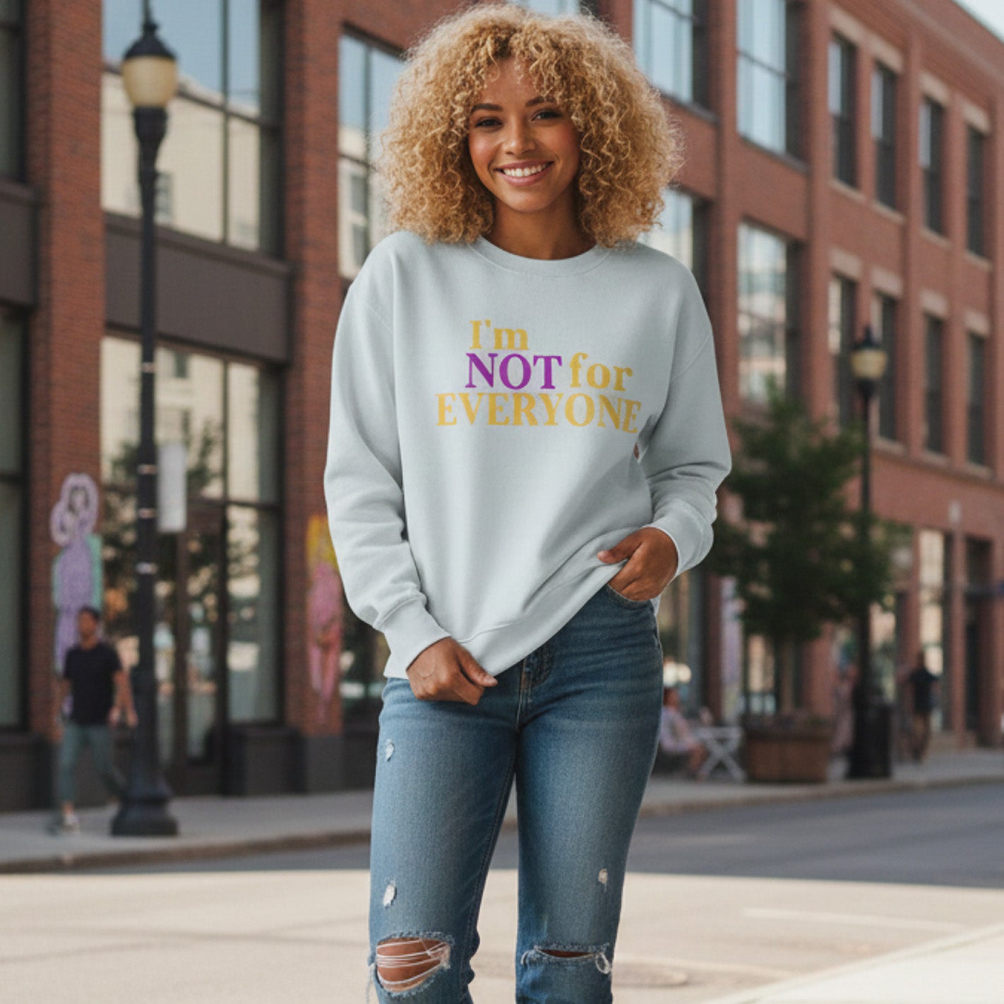 A smiling woman with curly blonde hair wears a light sweatshirt that says "I'm NOT for EVERYONE" in purple and yellow letters, standing on a city sidewalk with brick buildings and a streetlamp in the background.