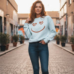 A woman stands in the middle of a cobblestone street, wearing a light blue sweatshirt with a smiley face and heart eyes, and blue jeans. She is smiling and surrounded by urban buildings and potted plants.