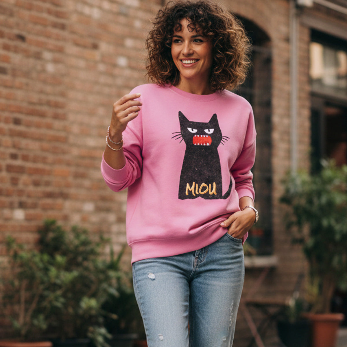 Woman wearing a pink sweatshirt with a black cat design and 'MIOU' text, standing in front of a brick wall.
