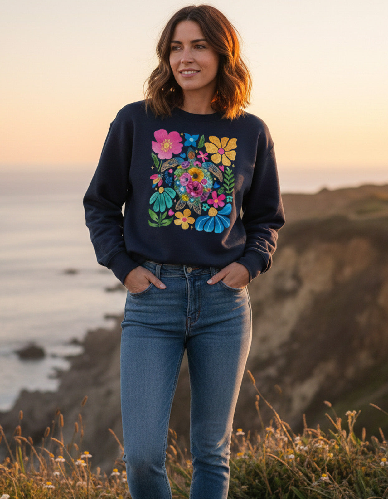 Woman wearing a dark blue sweatshirt with a colorful floral design, standing on a cliff overlooking the ocean at sunset.
