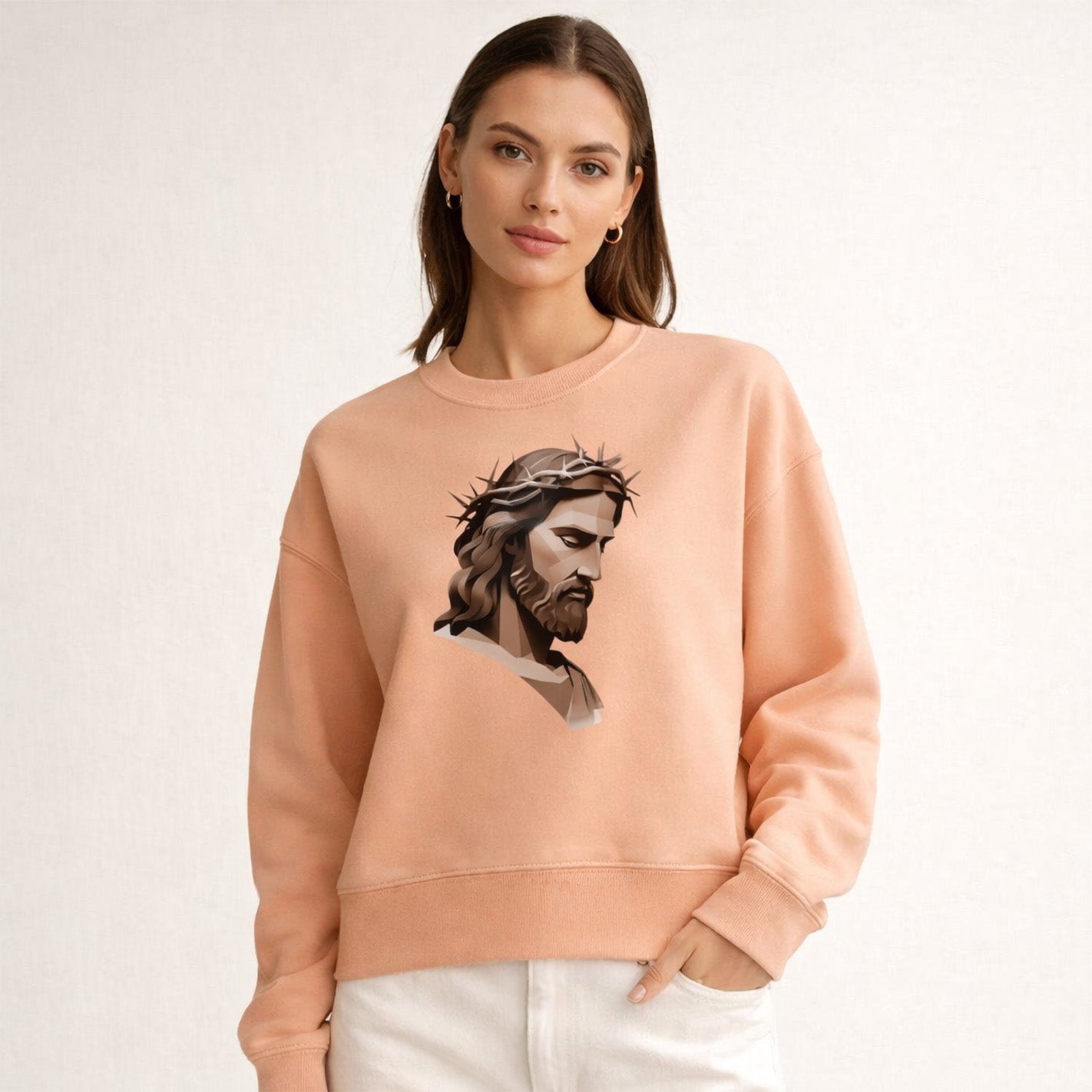 A woman wearing a peach-colored sweatshirt with an illustration of Jesus Christ crowned with thorns printed on the front stands against a plain light background.